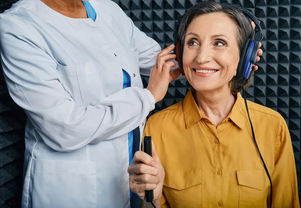 A senior woman smiling while a hearing clinic specialist places over the ear headphones on her to take a hearing test in a soundproof booth during a hearing aid consultation.